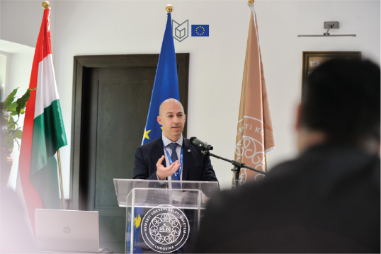 Man in front of hungarian, european and Ludovika University flag, giving a speech. Wears blue tye and dark suit. Gesticulating with his hand.