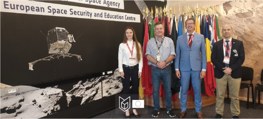 A group of four people standing before european flags and next to a drop-back of an Space Capsle landing on the moon.