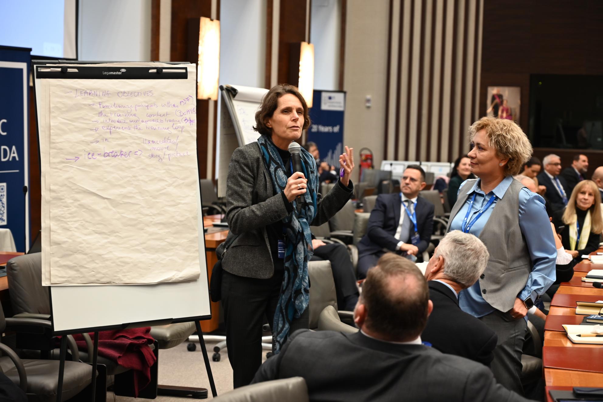 A woman with a microphone and a whiteboard presents to a group of people. 