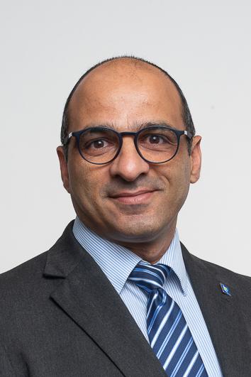 Christoforos Christoforou in a dark suit. Light blue shirt, blue striped tie. Wears glasses