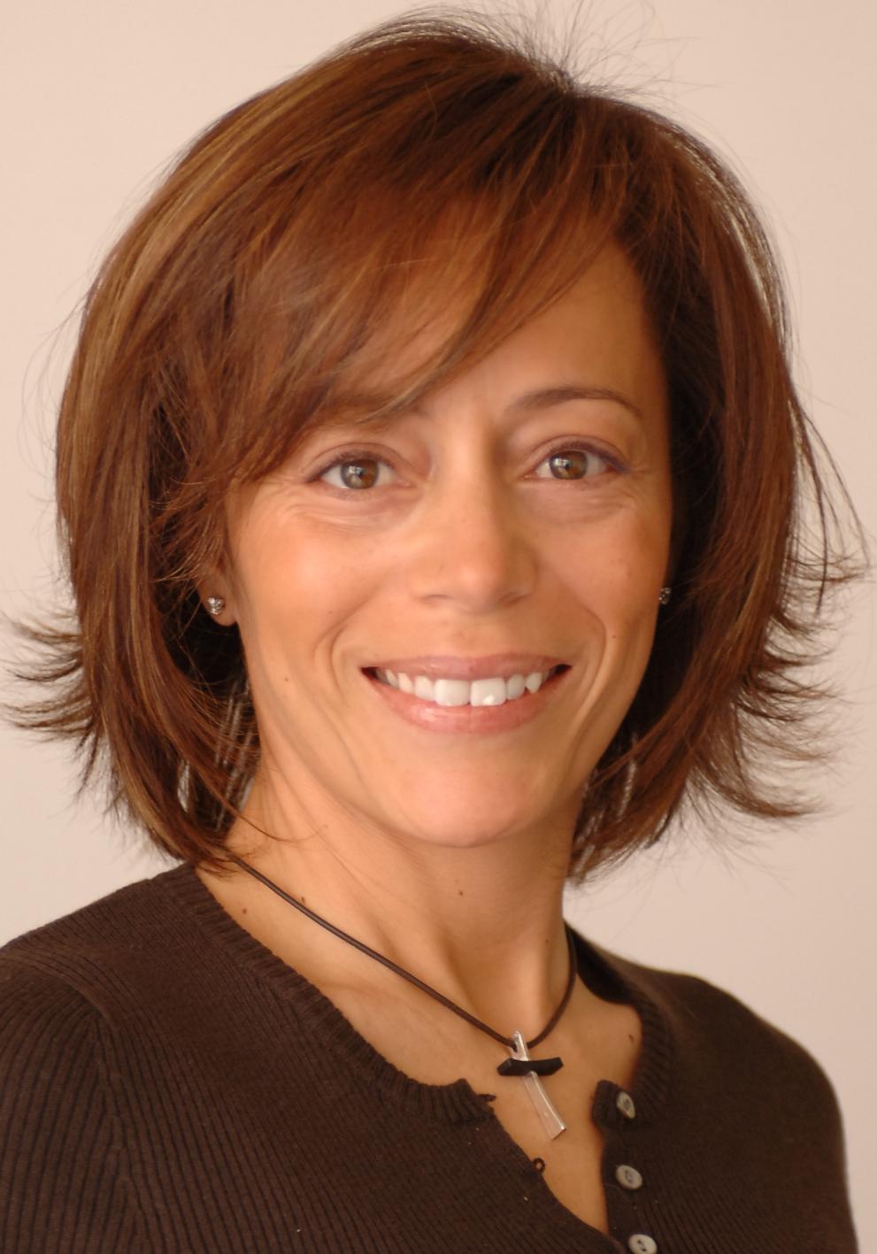 Maria Penedos. Brown, medium long hair, brown eyes. Smiling. Wearing abrown top and a chain with a cross.