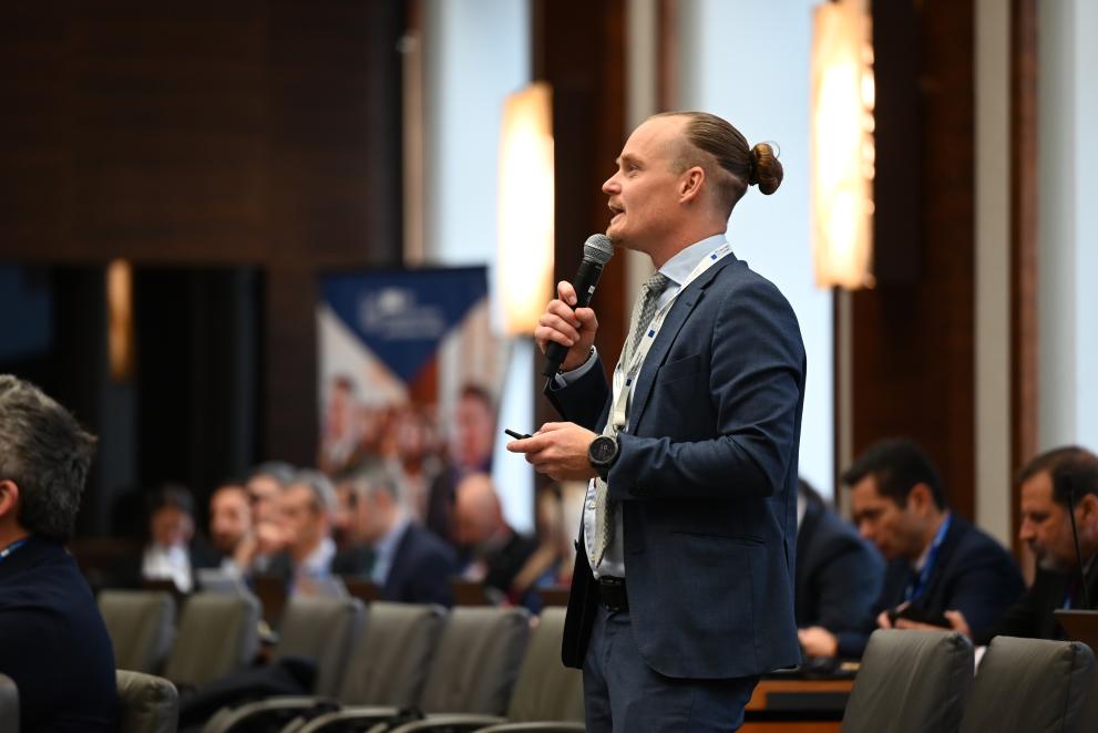 A man holding a microphone and talking. Blue suit, long hair in a bun. 