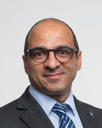 Christoforos Christoforou in a dark suit, blue shirt, blue striped tie. Brown glasses. 