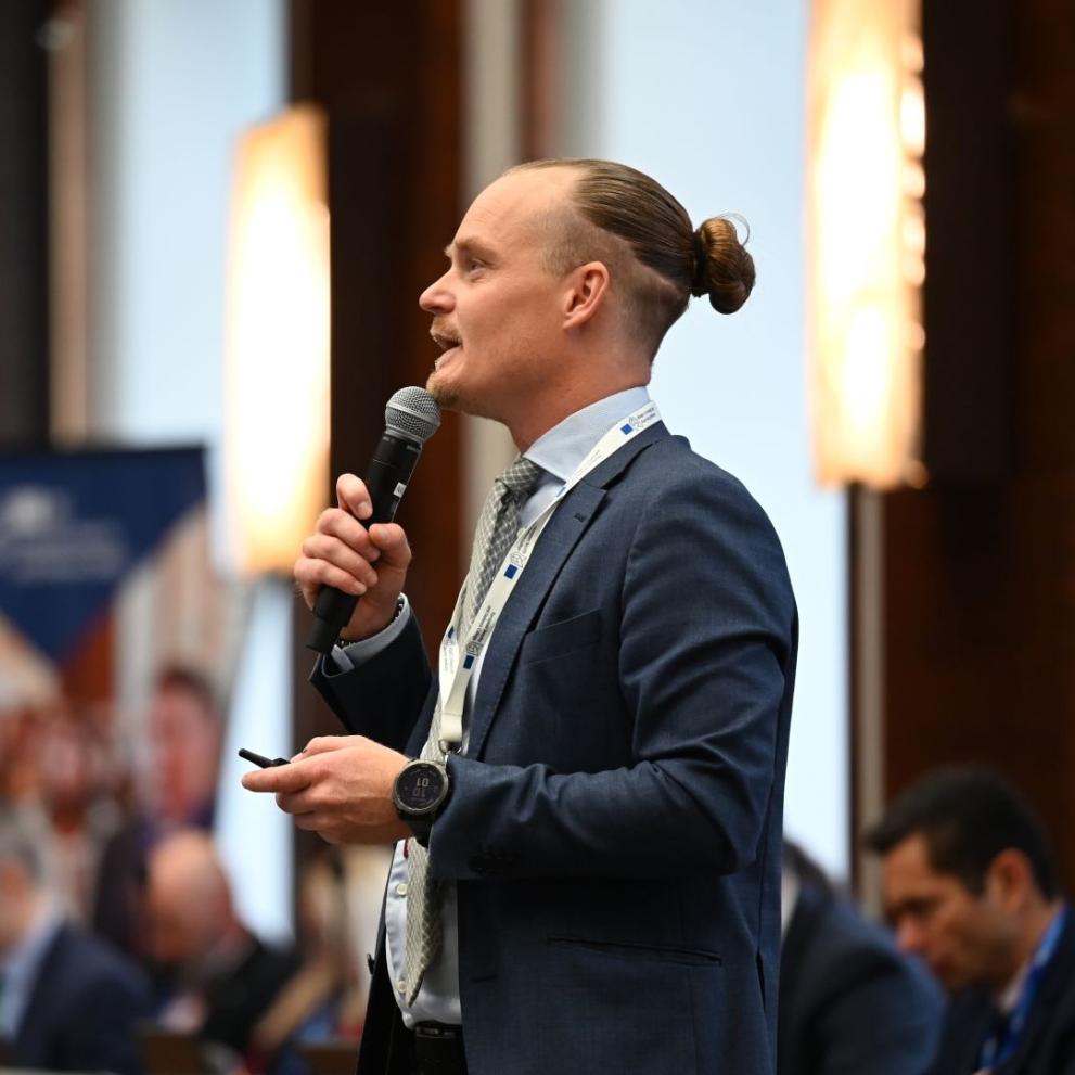 Isak Enstrom with a microphone, speaking during a conference. He is wearing a blue suit wih a tie.