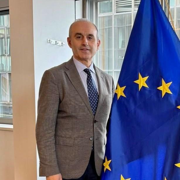 Dr Alin Bodescu next to an European Flag. He is wearing an grey jacket, light blue shirt and a blue tie. 