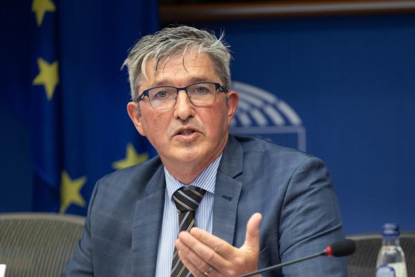 Picture of Fergal O Regan speaking in Parliament. In the background you can see a european flag and the symbol of the parliament. Only the upper half of Fergal O Regen is visible as he sits behind a desk.