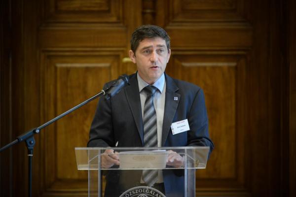 Dirk Dubois giving a speech in front of a wodden wall. He stands behind a lectern. He is wearing a dark suit and a tie.