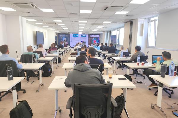 Group of participants sitting on single desk and focusing on a lecture in front. Approx. 25 people are sittingon the desk. 